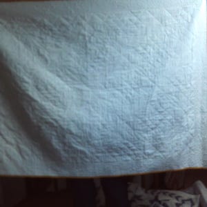 May include: A light blue quilt with a diamond pattern. The quilt has a yellow border. The quilt is hanging in a room. The quilt is made of a soft material.