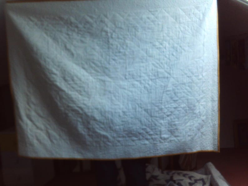 May include: A light blue quilt with a diamond pattern. The quilt has a yellow border. The quilt is hanging in a room. The quilt is made of a soft material.