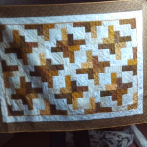 May include: A handmade quilt with a geometric pattern. The quilt features a white centre with a repeating pattern of brown and tan blocks. It has a brown border and a white inner border. The quilt is likely used for bedding or decoration.