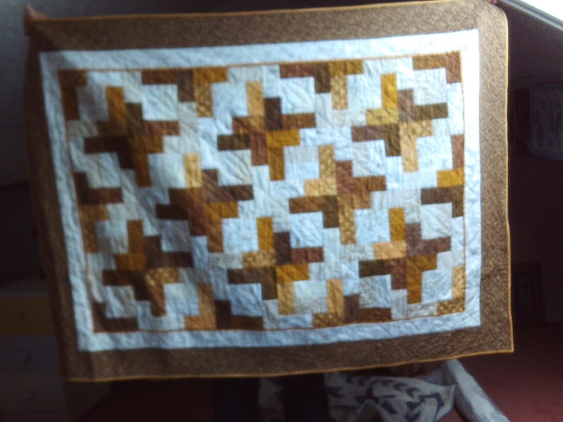 May include: A handmade quilt with a geometric pattern. The quilt features a white centre with a repeating pattern of brown and tan blocks. It has a brown border and a white inner border. The quilt is likely used for bedding or decoration.