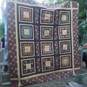 May include: A large, handmade quilt with a patchwork design. The quilt features a grid of squares with various patterns and colours, including red, blue, gold, and green. The border has a geometric pattern. The quilt is being held up outdoors.