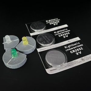 May include: Three small gray plastic bases with clear, yellow, and green LED lights mounted on top. The bases are next to a package of three CR2032 lithium batteries. The package has the text "Nightkonic Lithium Batteries CR2032 3V" printed on it.