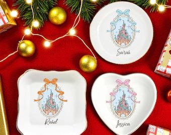 May include: Three white decorative trinket dishes with gold trim, each featuring a castle design and a name. One is round, one square, and one heart-shaped, set on a red surface with Christmas decorations.