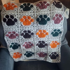 May include: A crocheted blanket featuring a grid pattern with colorful paw prints. The paw prints are in shades of orange, dark green, burgundy, and dark purple, set against a white background. The blanket is likely handmade.