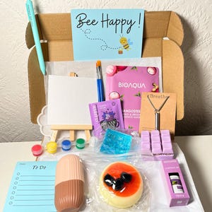 May include: A brown box filled with various items, including a small easel, paint, a face mask, a necklace, a to-do list, and a small cake. The box has a blue and white card on top that says "Bee Happy!"