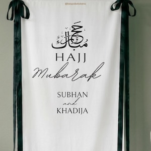 Op de afbeelding: Een witte stoffen wandhanger met zwarte tekst die Hajj Mubarak leest, gevolgd door de namen Subhan en Khadija. De banner is voorzien van zwarte linten aan de bovenhoeken en verticale zwarte lintafwerking langs de zijkanten.