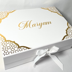 May include: A white gift box featuring gold geometric patterns on the corners and around the centre. The name "Maryam" is written in gold script. A white ribbon is tied in a bow on the front.