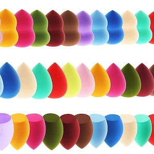 Op de afbeelding: Een verzameling kleurrijke make-upsponzen in verschillende vormen en tinten, waaronder roze, geel, groen, bruin, paars, blauw en oranje. De sponzen zijn in nette rijen gerangschikt tegen een witte achtergrond.
