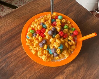 May include: A close-up of a bowl of cereal with milk. The cereal is a mix of small, square-shaped pieces and round, colorful pieces. The bowl is orange and there is a silver spoon sticking out of the cereal.