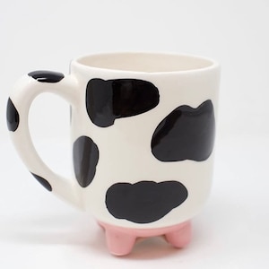 Może przedstawiać: Biały ceramiczny kubek z czarnymi plamami krowy i różową podstawą w kształcie kopyt. Uchwyt również ma plamy. Zabawny design na filiżankę kawy lub herbaty.