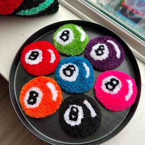 May include: A collection of seven colorful, handmade, round coasters resembling billiard balls, each with a white number eight design. The coasters are in various colors, including red, green, purple, orange, blue, black, and pink. They are arranged on a black tray.