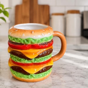 May include: A ceramic mug shaped like a hamburger, featuring layers of a bun, lettuce, tomato, cheese, and a patty. The mug has a brown handle and a white interior. The mug is on a marble countertop.