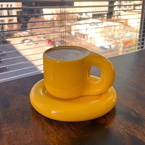 Puede incluir: Un juego de taza y platillo de café amarillo brillante. La taza tiene un diseño de asa único y se asienta sobre un platillo a juego. El juego es de cerámica y está colocado sobre una superficie de madera oscura.