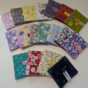 Könnte beinhalten: Eine Sammlung von Stoff-Nadelmappen mit verschiedenen floralen Mustern. Jede quadratische Mappe hat einen Knopfverschluss und ein elastisches Band oder Banddetail. Die Farben umfassen Rosa, Blau, Grün, Gelb und Rot.