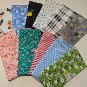 Könnte beinhalten: Eine Sammlung von Stoff-Buchhüllen in verschiedenen Mustern und Farben. Designs umfassen Blumen, Tupfen, Streifen und Zeichentrickfiguren. Jede Hülle ist rechteckig und hat einen Knopfverschluss.