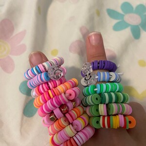May include: Two colorful beaded rings stacked on fingers. The rings are made of small, round, plastic beads in various colors, including pink, green, blue, yellow, and orange. The rings are stacked on top of each other, creating a colorful and eye-catching look.