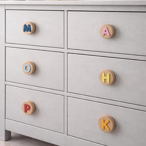 Könnte beinhalten: Eine hellgraue Kommode mit sechs Schubladen, jede mit einem Holzknauf mit einem farbigen Buchstaben. Die Buchstaben sind blau, rosa, orange, gelb und rot. Ein Teddybär und Holzhäuser stehen oben drauf.