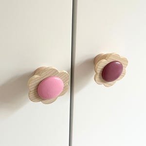 Może przedstawiać: Dwa uchwyty do szafek w kształcie kwiatów. Gałki mają jasną, drewnianą, karbowaną krawędź i gładki, owalny środek. Jedna gałka ma różowy środek, a druga środek w kolorze malwy. Gałki są zamontowane na białych drzwiach szafy.