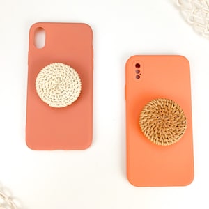 Puede incluir: Dos fundas de teléfono de color coral con agarres de teléfono tejidos diferentes. Un agarre es blanco y el otro es marrón.