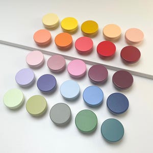 Può includere: Una collezione di ventuno pomelli rotondi per mobili in vari colori, tra cui giallo, arancione, rosa, rosso, viola, blu e verde. I pomelli sono disposti in file e colonne, mostrando una gamma di tonalità pastello e vivaci.