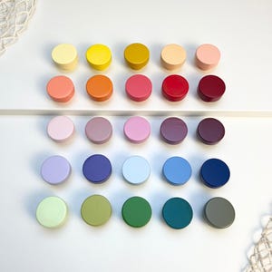 May include: A collection of twenty colorful, round cabinet knobs in various shades, including yellow, orange, pink, purple, blue, and green. The knobs are arranged in a grid pattern on a white surface, offering a range of color options for home decor projects.
