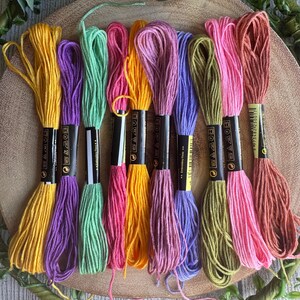 May include: A selection of embroidery threads in various colours, including yellow, purple, green, pink, orange, and brown. The threads are arranged on a wooden surface, each skein labelled. The image displays a range of bright colours, suitable for craft projects.