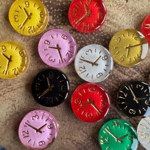 May include: A selection of small, circular clock faces in diverse colours, such as yellow, red, pink, and green. Each clock face features gold-coloured hands and numerals. The clocks are arranged in a random pattern.