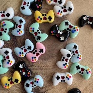 May include: Assortment of colourful miniature plastic game controllers in various colours, including black, white, yellow, pink, light blue and green. Each controller has a heart-shaped button and a plus sign button.