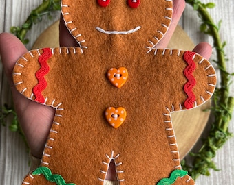 May include: A felt gingerbread man decoration with a stitched outline, red button eyes, and a smiling mouth. It features green and red decorative trim, and orange heart-shaped buttons. A jute string is attached for hanging.