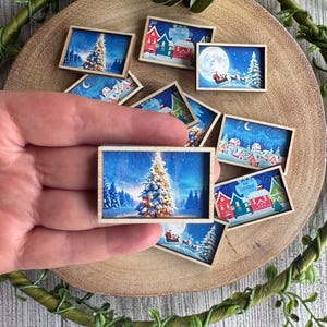 May include: A collection of miniature rectangular wooden frames containing printed Christmas scenes, including images of a decorated pine tree, a snowy village with colourful houses, and a silhouette of a sleigh flying across a full moon.