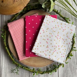 May include: Three fabric squares with different patterns. The first square is pink with white polka dots, the second square is red with white polka dots, and the third square is white with pink and gold polka dots.