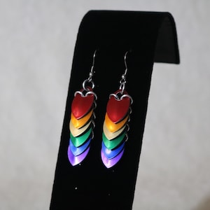 May include: Dangle earrings in a rainbow design. Each earring has a series of metal scales in red, orange, yellow, green, blue, and purple. The earrings are displayed on a black stand.