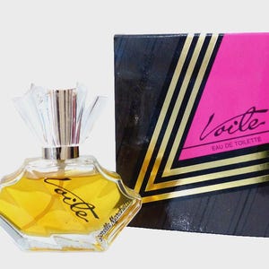 Può includere: Un flacone di profumo in vetro con liquido giallo, dotato di un tappo trasparente a forma di ventaglio e la scritta Voile Sorelle Marchelle. Accanto si trova una scatola rettangolare con un design nero e rosa acceso, strisce dorate e la scritta Voile Eau de Toilette.