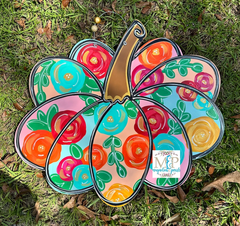 May include: A pink and blue pumpkin with a floral design. The pumpkin is painted with bright colors and has a black outline. The text "MP Magnolia Pearls" is in the bottom right corner.