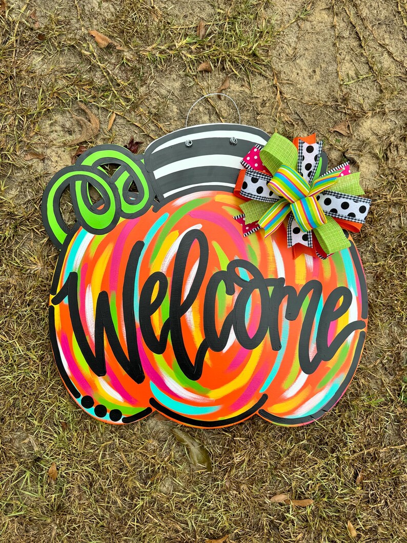 Puede incluir: Un letrero de bienvenida de madera con forma de calabaza con un dise&ntilde;o colorido y arremolinado en naranja, rosa, amarillo y turquesa. La palabra "Welcome" est&aacute; escrita en negro. La parte superior es a rayas blancas y negras, con un remolino verde y negro y un lazo decorativo.