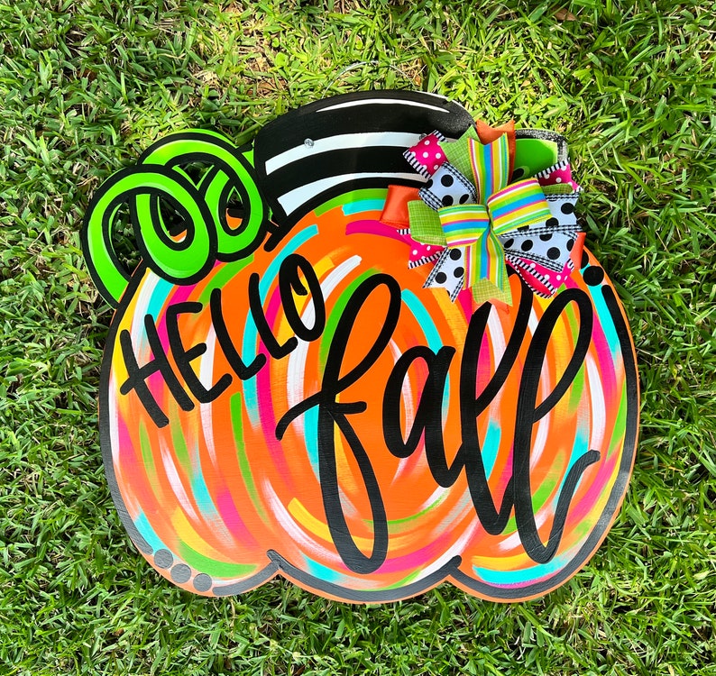 Puede incluir: Un letrero de madera con forma de calabaza con las palabras "HELLO fall" en escritura negra. La calabaza est&aacute; pintada de naranja con pinceladas coloridas y un tallo a rayas blanco y negro. Se adjunta un lazo decorativo con lunares y rayas.