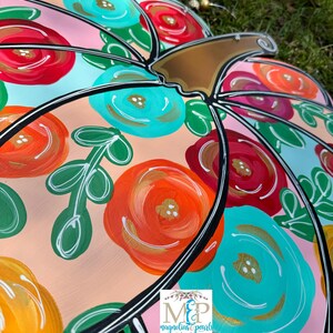 May include: A pink pumpkin door hanger with a black outline and a brown stem. The pumpkin is decorated with colorful flowers in shades of red, orange, yellow, blue, and green.