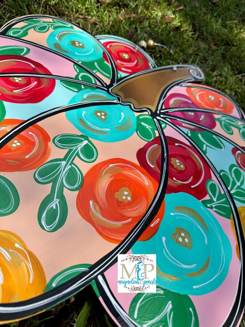 May include: A pink pumpkin door hanger with a black outline and a brown stem. The pumpkin is decorated with colorful flowers in shades of red, orange, yellow, blue, and green.
