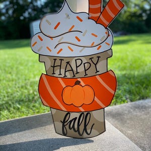 May include: A wooden door hanger shaped like a coffee cup with whipped cream and sprinkles. The cup has an orange and white striped band around it and a pumpkin on the front. The text "Happy fall" is written on the cup.