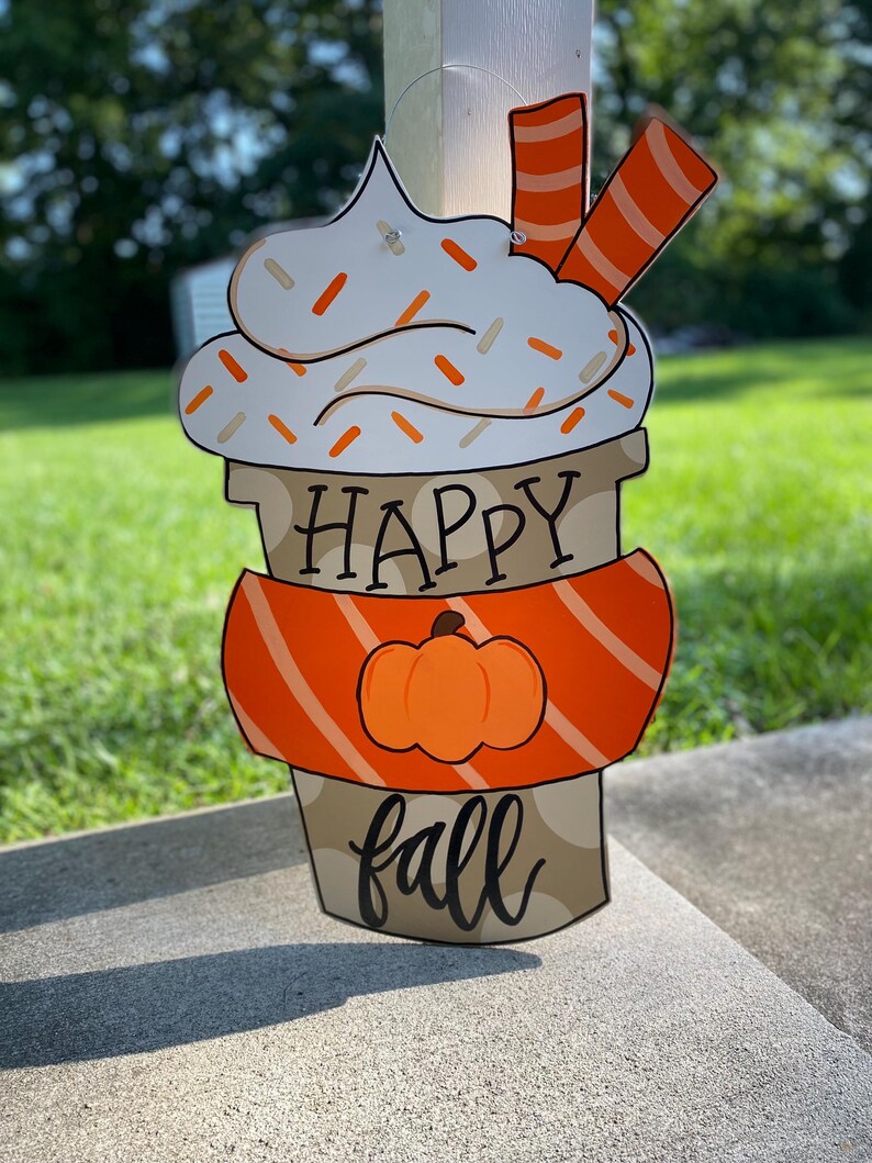 May include: A wooden door hanger shaped like a coffee cup with whipped cream and sprinkles. The cup has an orange and white striped band around it and a pumpkin on the front. The text "Happy fall" is written on the cup.