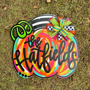Puede incluir: Un letrero de madera con forma de calabaza colorida con el texto "the Hatfields" en escritura negra. La calabaza tiene una parte superior a rayas blancas y negras, zarcillos verde ne&oacute;n y un dise&ntilde;o de remolino multicolor. Se adjunta un lazo decorativo.