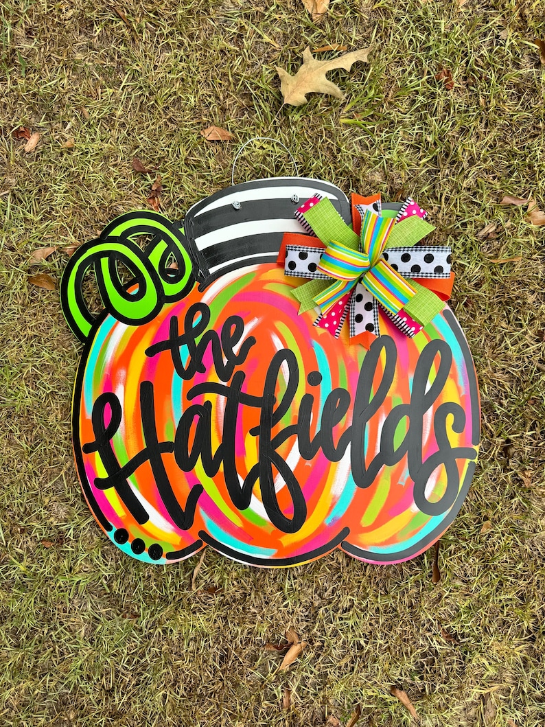 Puede incluir: Un letrero de madera con forma de calabaza colorida con el texto "the Hatfields" en escritura negra. La calabaza tiene una parte superior a rayas blancas y negras, zarcillos verde ne&oacute;n y un dise&ntilde;o de remolino multicolor. Se adjunta un lazo decorativo.