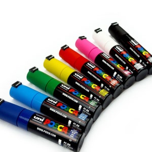 May include: A set of ten Uni Posca paint markers in various colors, including black, white, red, pink, yellow, green, blue, and light blue. The markers are arranged in a row, with the black marker at the top and the light blue marker at the bottom. Each marker has the Uni Posca logo and website address printed on the side. The markers are all labeled with the color name and a PC-8K code.