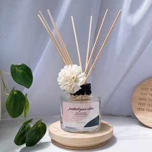 Peut inclure: Une bouteille de diffuseur en verre transparent avec une étiquette noire et blanche qui indique "protect your vibes" et "Black Toumaline". La bouteille est remplie d'un liquide clair et des bâtonnets en bois en sortent. Une fleur blanche est posée sur les bâtonnets. La bouteille est posée sur une base en bois.