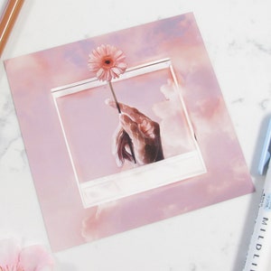 May include: A pink flower is held by a hand in a white frame against a pink and white cloudy background.