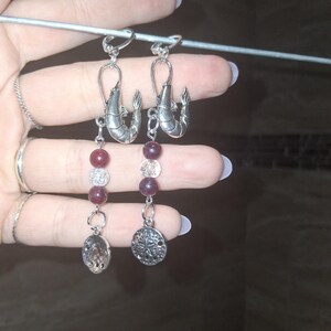 May include: A pair of dangle earrings featuring silver-toned shrimp charms at the top. Each earring has a strand of dark red beads, clear beads, and a silver-toned sand dollar charm. The earrings are suspended from a silver hook.