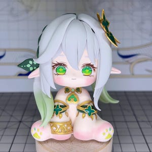 Puede incluir: Figura artesanal de un personaje con cabello blanco, ojos verdes y orejas puntiagudas. La figura tiene un tono de piel rosa claro y está adornada con detalles verdes y dorados. Está posada sentada sobre una pequeña base de madera redonda.