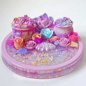 May include: A pink and purple resin trinket dish with a seashell, flowers, and two small decorative boxes. The dish is filled with glitter and the words "love you" are written in gold lettering.