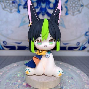 Puede incluir: Figura artesanal de un personaje con cabello negro y verde, orejas grandes con interiores rosas y ojos verdes. La figura lleva una capa azul con detalles en naranja y un collar de flores amarillas. La figura está sentada sobre una base de madera.