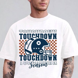 May include: White t-shirt with a blue and brown graphic of a football helmet and the text "Touchdown Season".